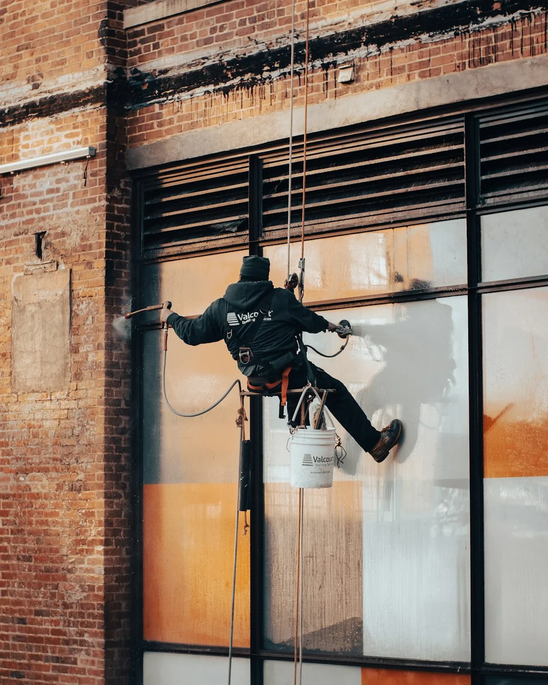 Black and white from below of man steeplejack in protective helmet with professional equipment cleaning glass wall of modern tower